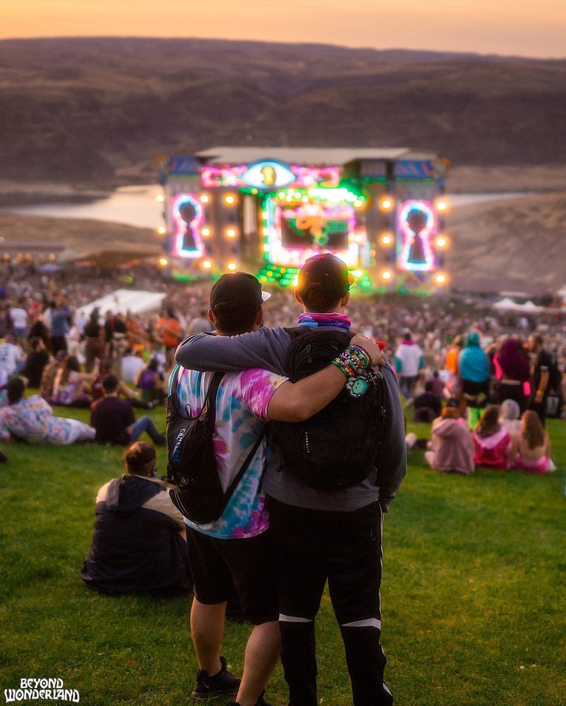 Two people looking down a crowded hill to a brightly lit stage, with a river and mountains behind it.