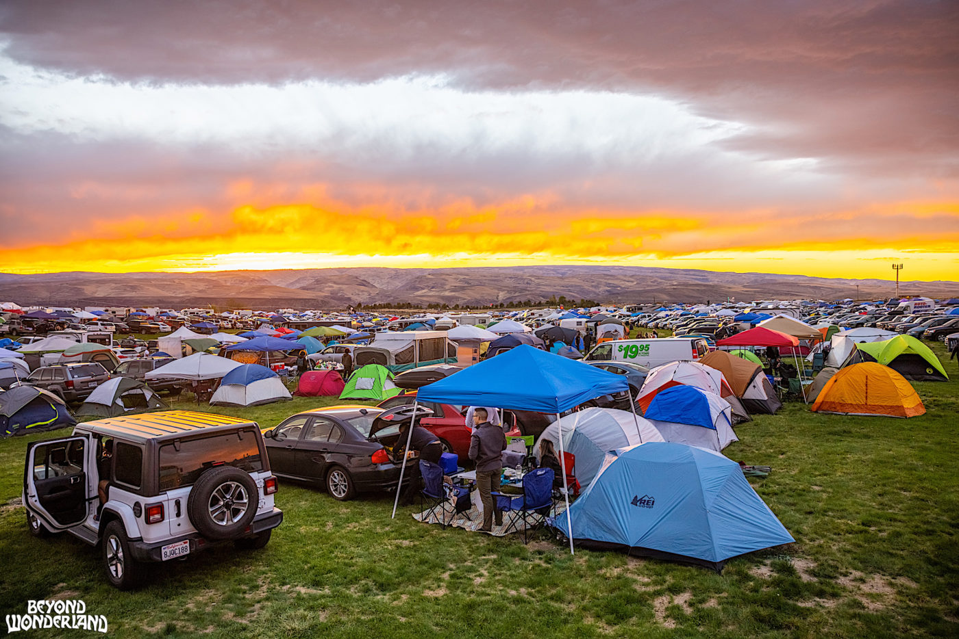A campground full of cars, canopies, and tents, with the sun setting in the distance.