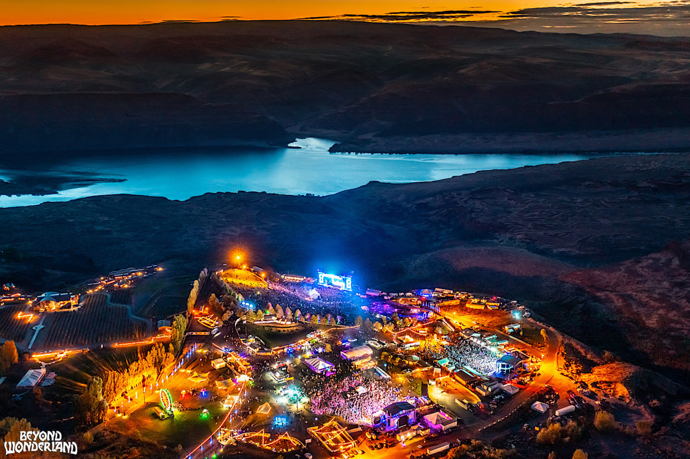 An wide overhead shot of a music festival at night. The sun sets behind a river in the distance.