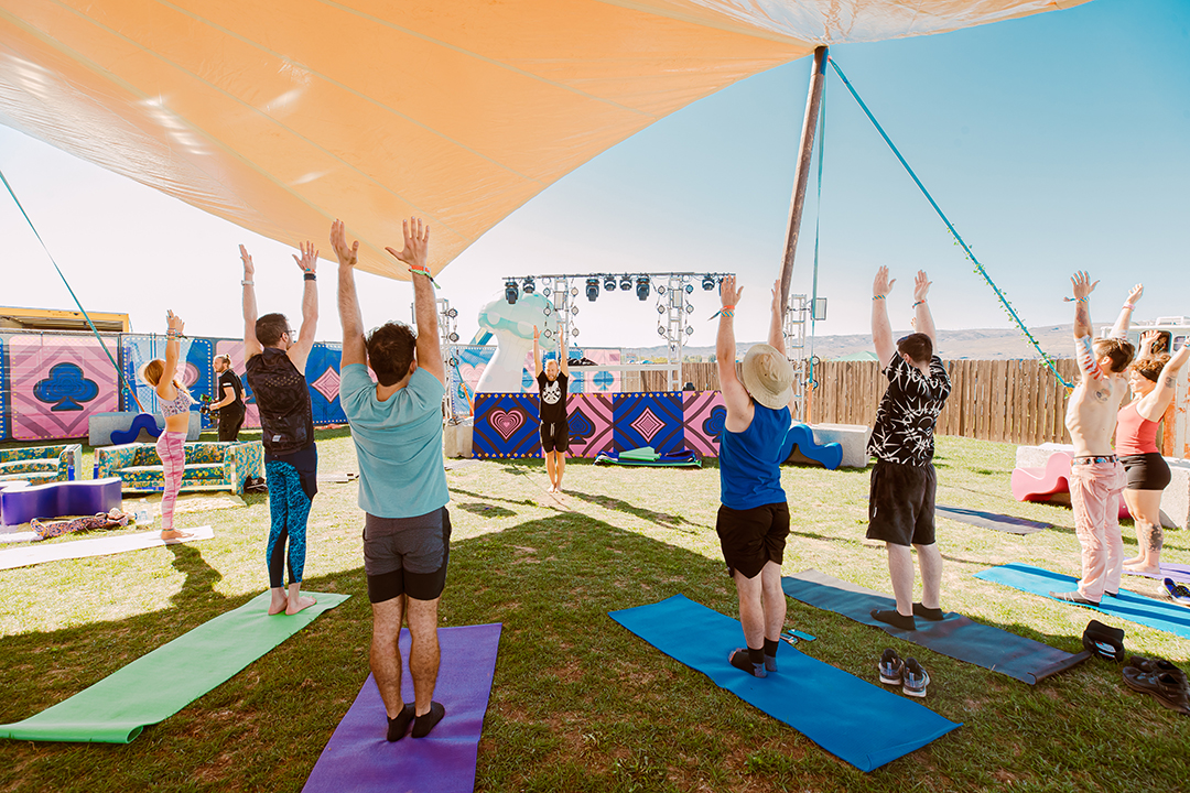 Eight people stretching upward as they do yoga in the shade under a white canopy.